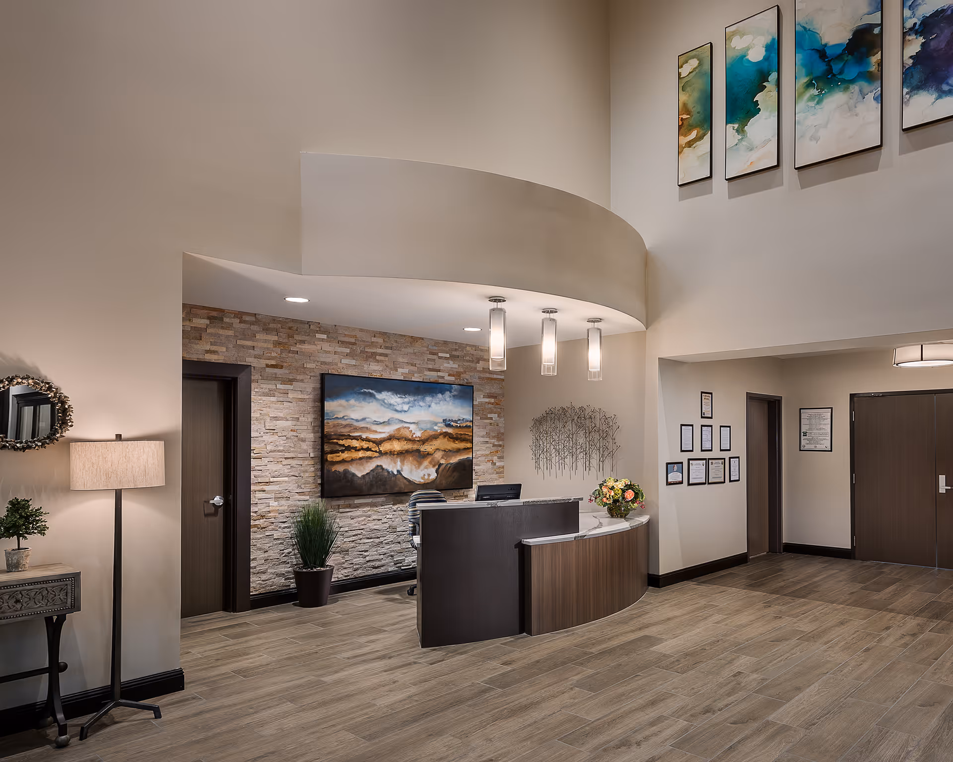 Reception area with a curved wooden desk, a computer, and a floral arrangement. Behind the desk is a stone accent wall with a large landscape painting and a potted plant. The floor is wood-style tile, and the walls are painted beige. There are three pendant lights hanging from the ceiling above the desk, and several framed certificates and artwork on the walls. A floor lamp and small table with a plant are on the left side.