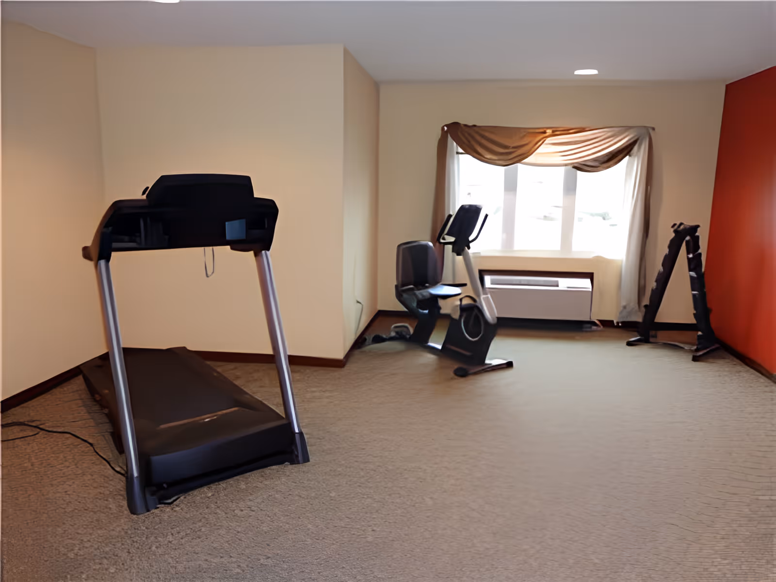 A small exercise room with a treadmill on the left, a recumbent stationary bike near a window with beige curtains, and a rack of dumbbells against a red accent wall. The room has beige walls and carpeted floor.