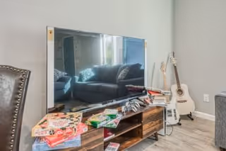 A modern living room area featuring a large flat-screen TV on a wooden media console with various board games and video game controllers on top. In the background, there is a gray sofa with cushions and two guitars resting against the wall on the right side.