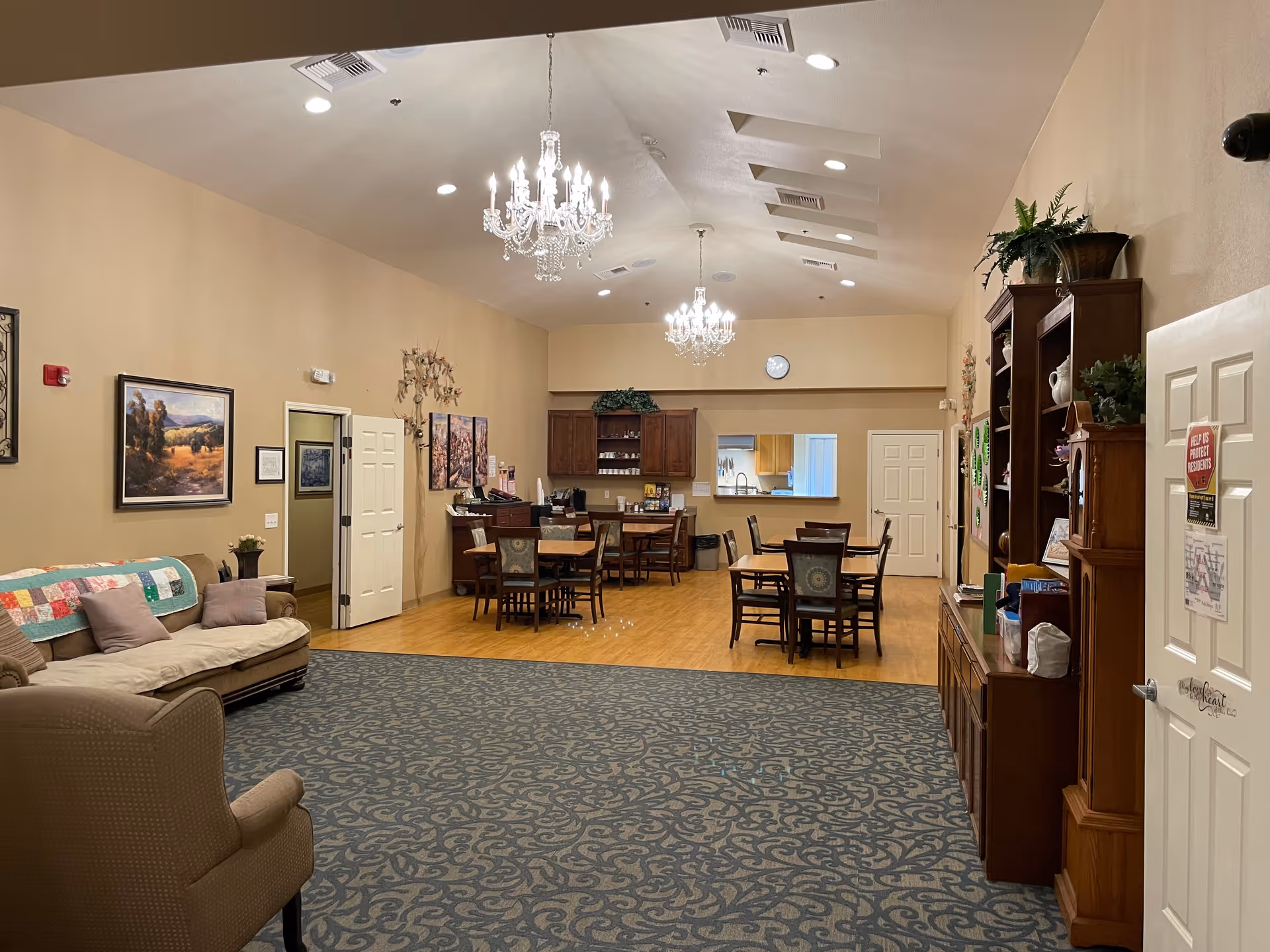 Spacious senior living common area with sofas and armchairs in the foreground, dining tables and chairs near a serving kitchen, and chandeliers overhead.