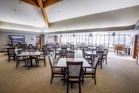 A spacious dining room with multiple square tables and cushioned chairs arranged neatly. The room features large windows along one wall allowing natural light to fill the space. There is a TV mounted on the wall, a stone accent wall, and a wooden ceiling beam visible. The carpeted floor and neutral color palette create a warm and inviting atmosphere.