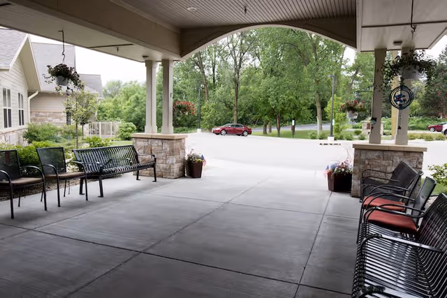 Covered outdoor seating area with metal benches and chairs with red cushions, stone pillars, hanging flower baskets, and a view of a parking lot with trees and a red car in the background.