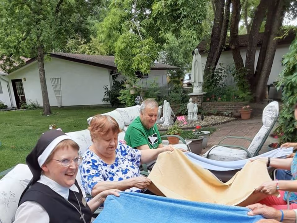 A group of elderly people and a nun sitting outdoors on cushioned chairs and sofas in a garden area, holding colorful blankets together. The background shows green trees, a white building, and religious statues surrounded by plants and flowers.
