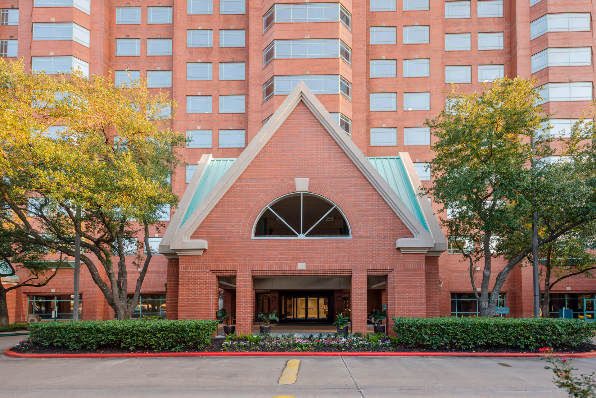 Entrance of a red-brick multi-story building with a triangular portico, flanked by trees and landscaped shrubs.