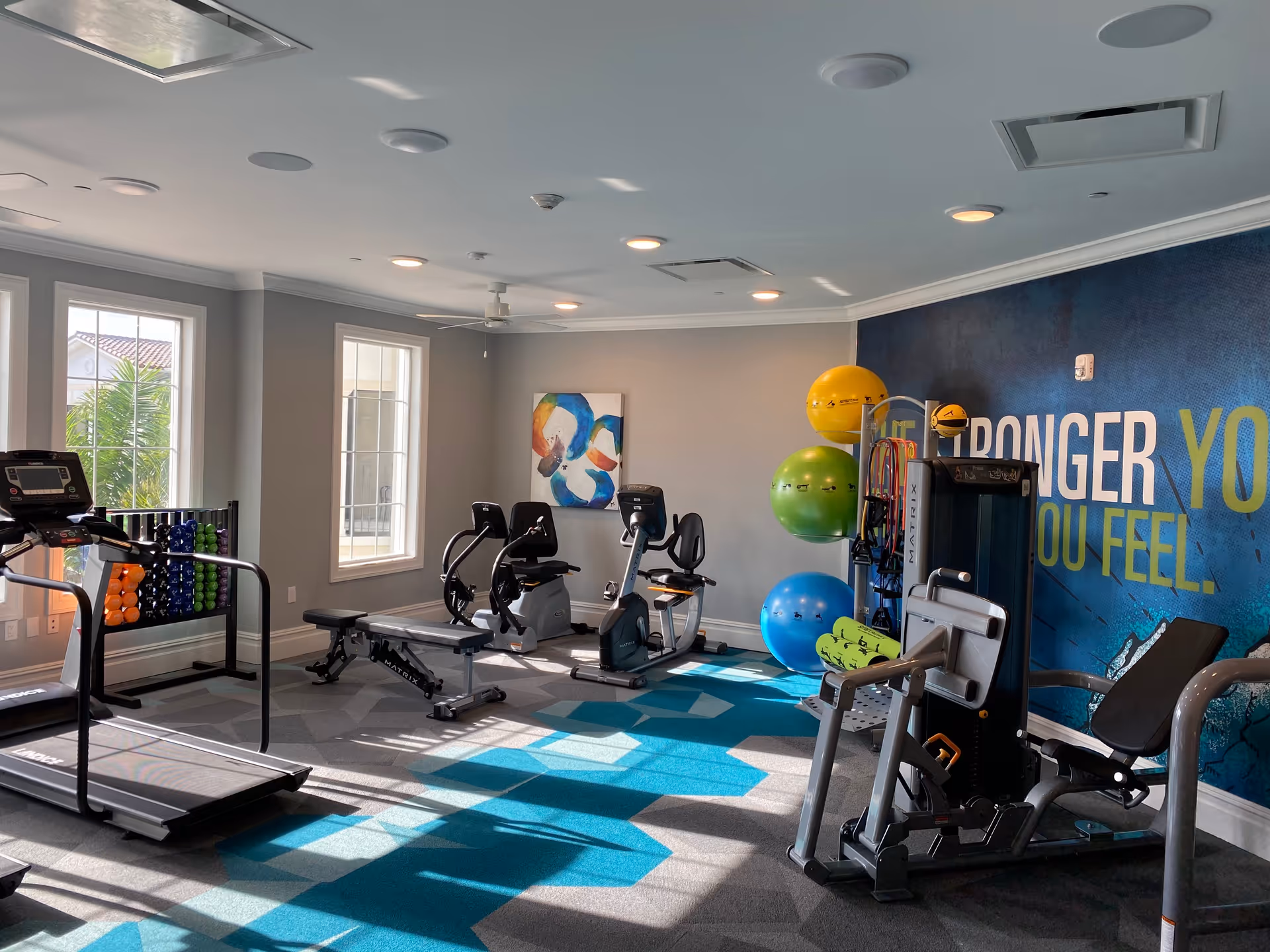 A well-lit fitness room with exercise equipment including a treadmill, stationary bikes, weight bench, and strength training machines. Colorful exercise balls and dumbbells are neatly arranged. The room has large windows letting in natural light and a blue wall with motivational text partially visible.