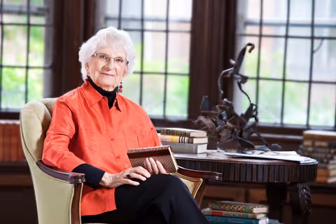 An elderly woman with white hair and glasses sits in a green upholstered armchair holding a book. She is wearing a bright orange shirt over a black top and is smiling gently. Behind her is a wooden table with several stacked books and a decorative sculpture, set in a room with large windows letting in natural light.