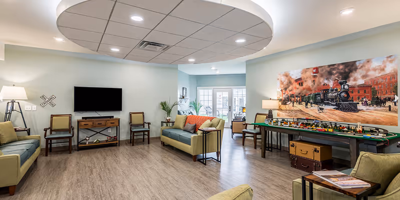 A spacious senior living common area with light green sofas, wooden chairs, a flat-screen TV mounted on the wall, and a large table with a model train set. The room has wood flooring, soft lighting from lamps, and a large painting of a vintage steam train on the wall.