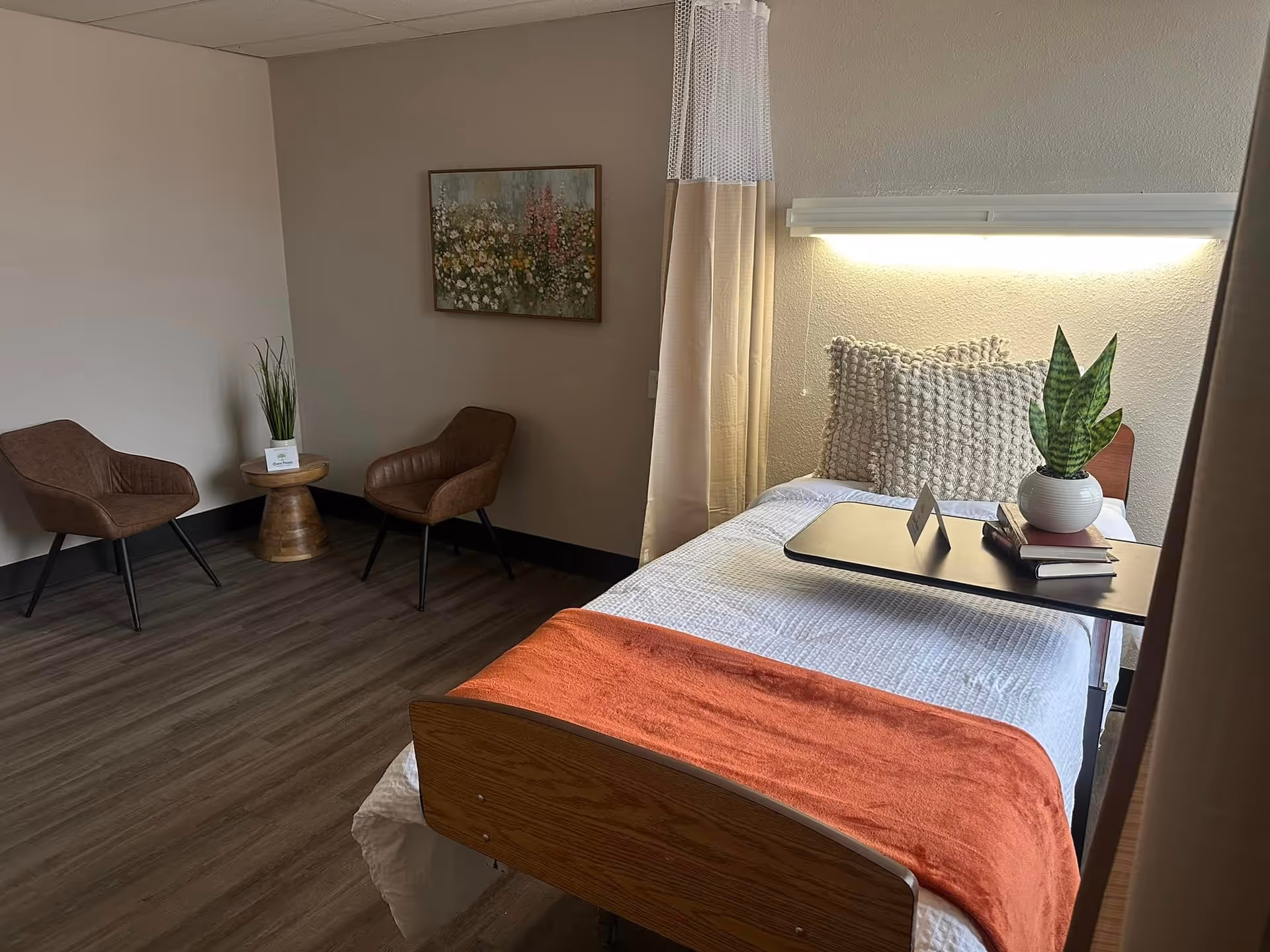 A cozy bedroom in a senior living facility with a single bed covered in white bedding and an orange throw blanket. The bed has two textured pillows and a small table on wheels with a potted plant and books. There are two brown chairs and a small round wooden table with a plant in the corner. A floral painting hangs on the wall above the chairs. The room has wood flooring and neutral-colored walls.