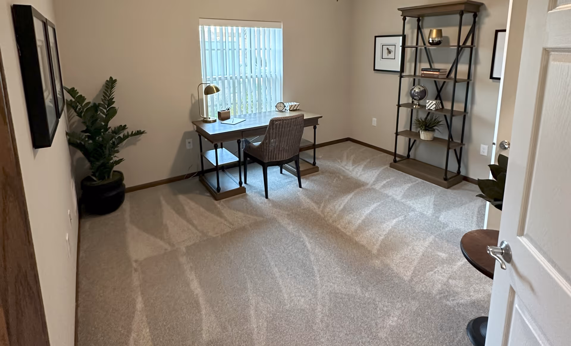 A bright room with beige walls and carpeted floor featuring a wooden desk with a chair placed in front of a window with vertical blinds. The desk has a gold lamp, a clock, and some small decorative items. To the right, there is a tall shelving unit with books, a globe, and plants. On the left side, there is a large potted plant and framed pictures on the wall.