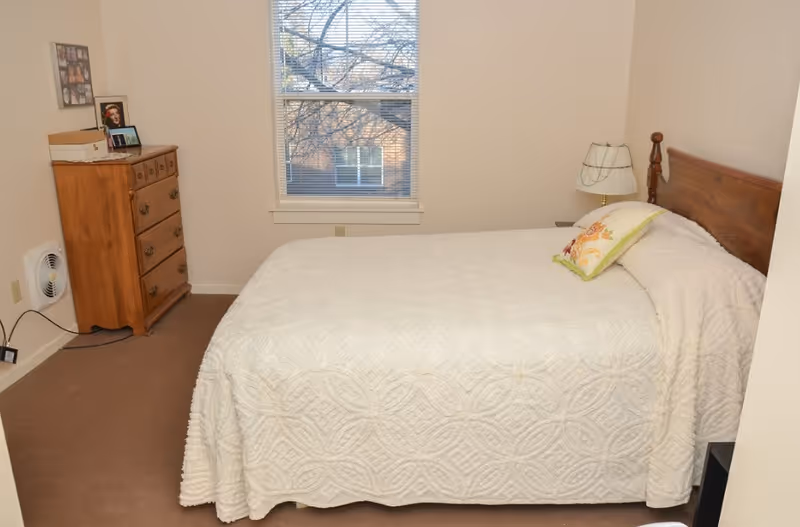 A simple bedroom with a bed covered in a white textured bedspread and a decorative pillow. There is a wooden headboard, a bedside table with a lamp, a wooden dresser with framed photos on top, and a window with blinds showing a view of a tree and a building outside.