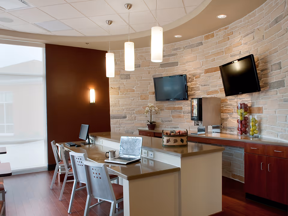 A modern interior room with a curved stone accent wall, two wall-mounted flat screen TVs, a countertop with a coffee machine and decorative fruit jars, and a long table with several chairs and laptops placed on it. The room has pendant lights hanging from the ceiling and a large window with a sheer curtain.