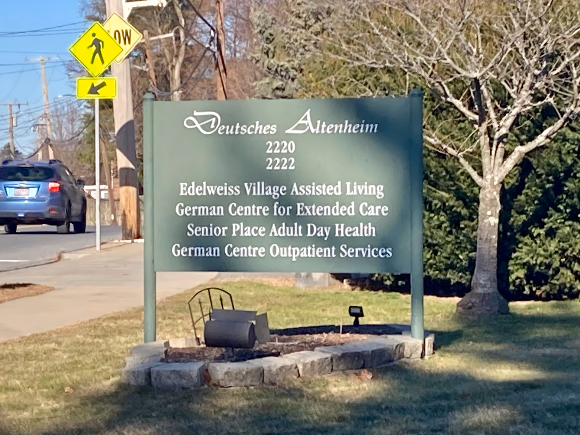 Outdoor view of a green signboard for Deutsches Altenheim with addresses 2220 and 2222. The sign lists services including Edelweiss Village Assisted Living, German Centre for Extended Care, Senior Place Adult Day Health, and German Centre Outpatient Services. The sign is placed on a grassy area with a tree and some bushes in the background. A pedestrian crossing sign and a blue car on the road are visible to the left.