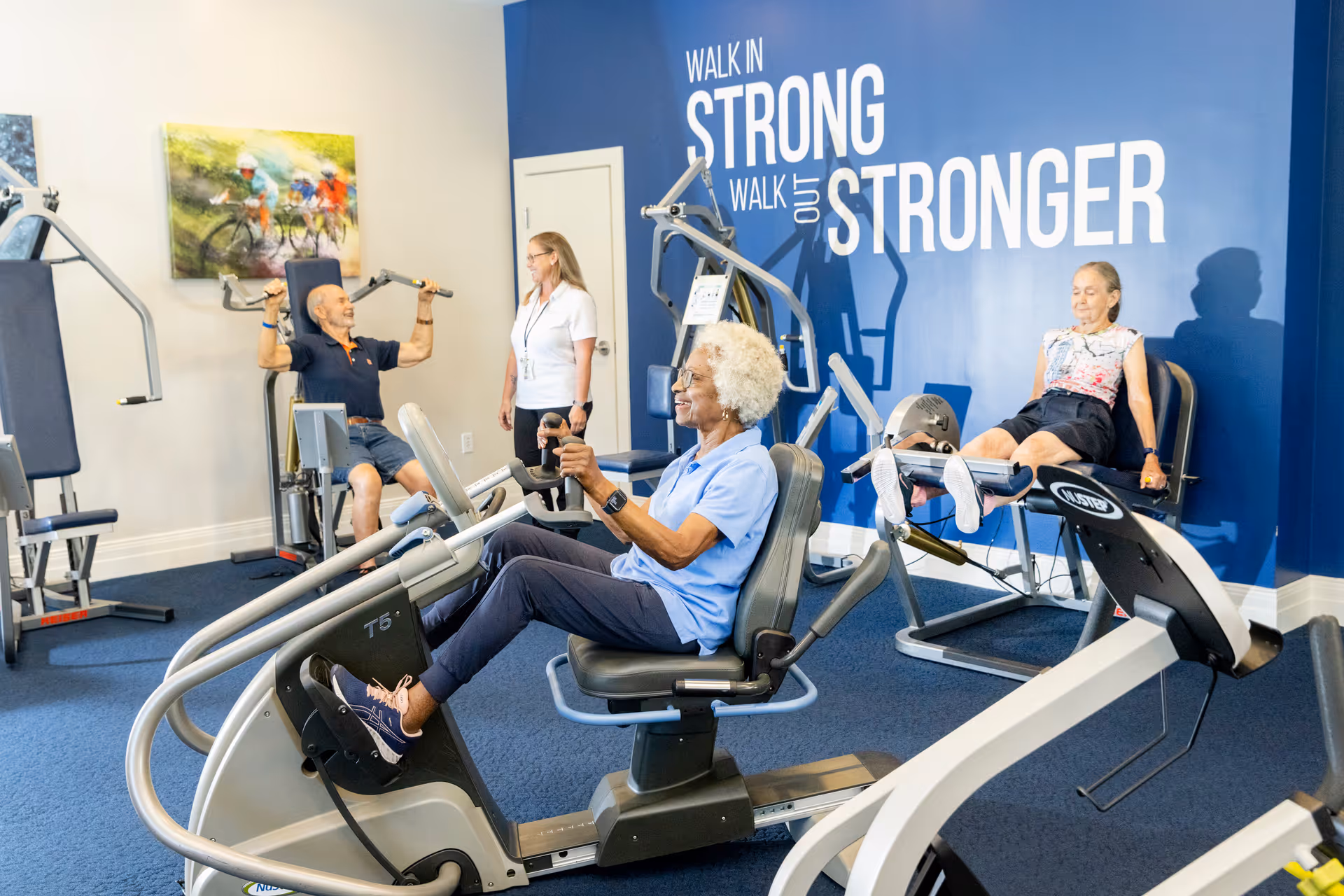 Three elderly individuals exercising on various gym equipment in a fitness room with a blue wall that has the motivational text 'Walk in Strong, Walk out Stronger'. A staff member is standing and observing them. The room has blue carpeting and a colorful painting on the wall.