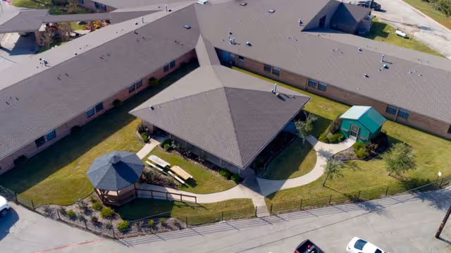 Aerial view of Southland Rehabilitation And Healthcare Center showing a large building with a central courtyard. The courtyard features a gazebo, benches, a small green shed, landscaped grass areas, and paved walkways. The building has a gray roof and brick walls, surrounded by a fence and adjacent to a parking area.