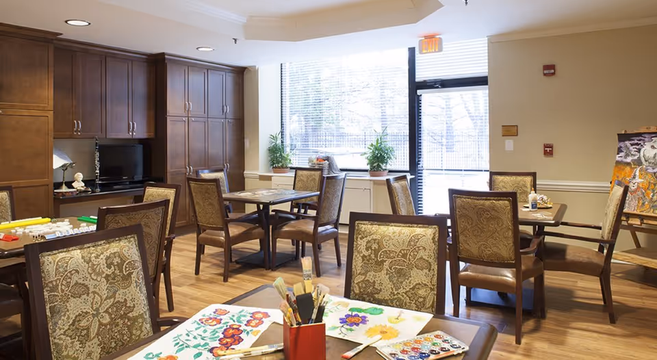 Bright communal activity room with several tables and upholstered chairs, art supplies on a foreground table, built-in cabinets, and a large window and exit door.