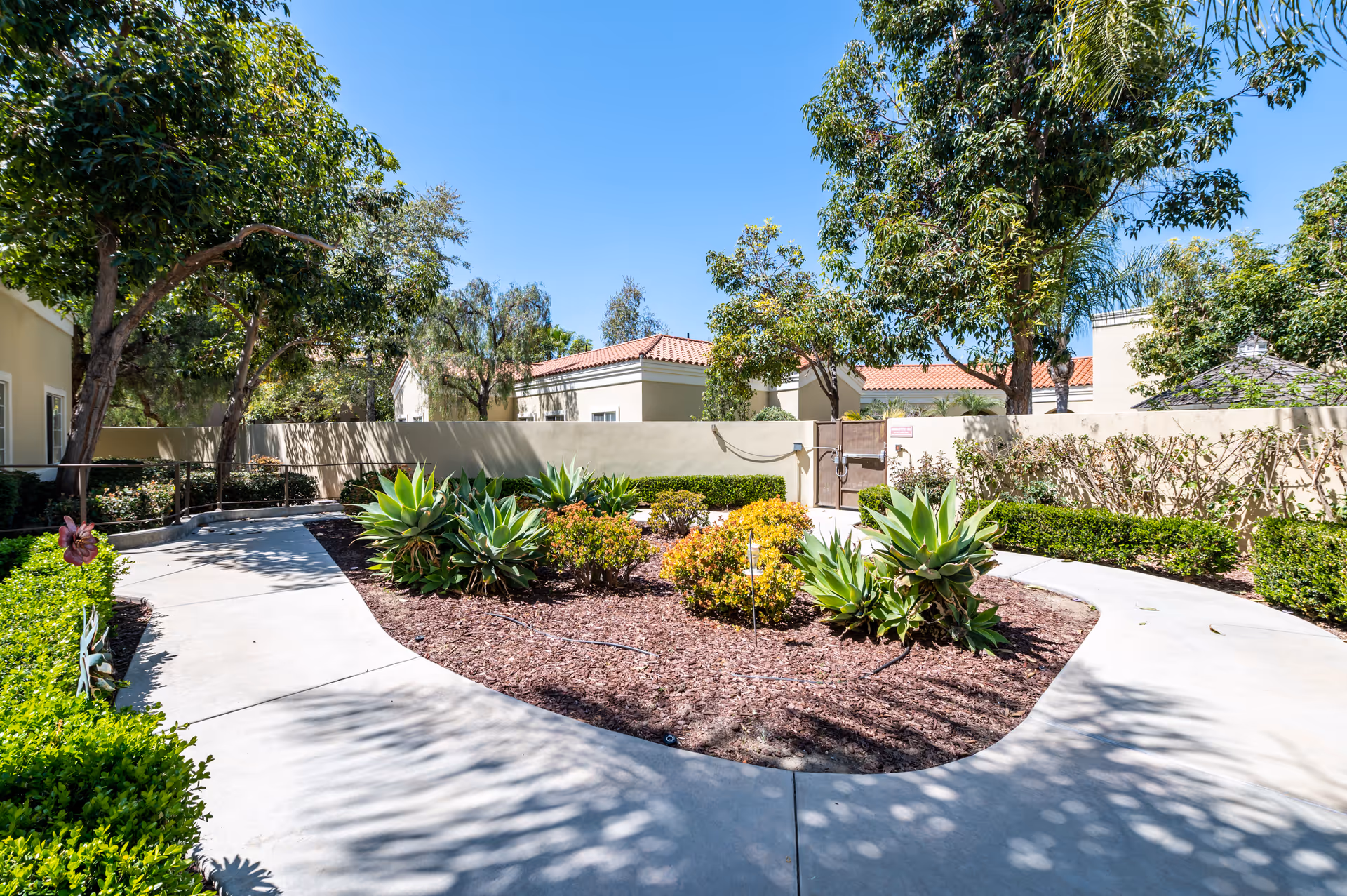 A sunny outdoor garden area at San Juan Hills Healthcare Center featuring a curved concrete walkway surrounding a landscaped bed with various green plants and shrubs. Trees provide partial shade, and beige buildings with red-tiled roofs are visible in the background under a clear blue sky.