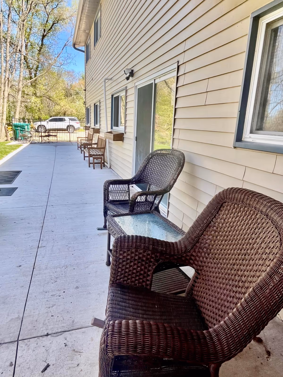Concrete patio alongside a beige-sided building with wicker chairs, a glass-topped table, and sliding doors.