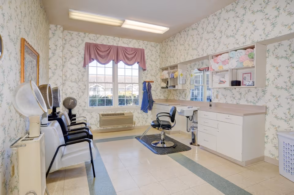 A bright room with floral wallpaper featuring a hair salon setup including three hair dryers on the left, a styling chair in front of a counter with a sink, shelves stocked with colorful towels, and a large window with a pink valance letting in natural light.