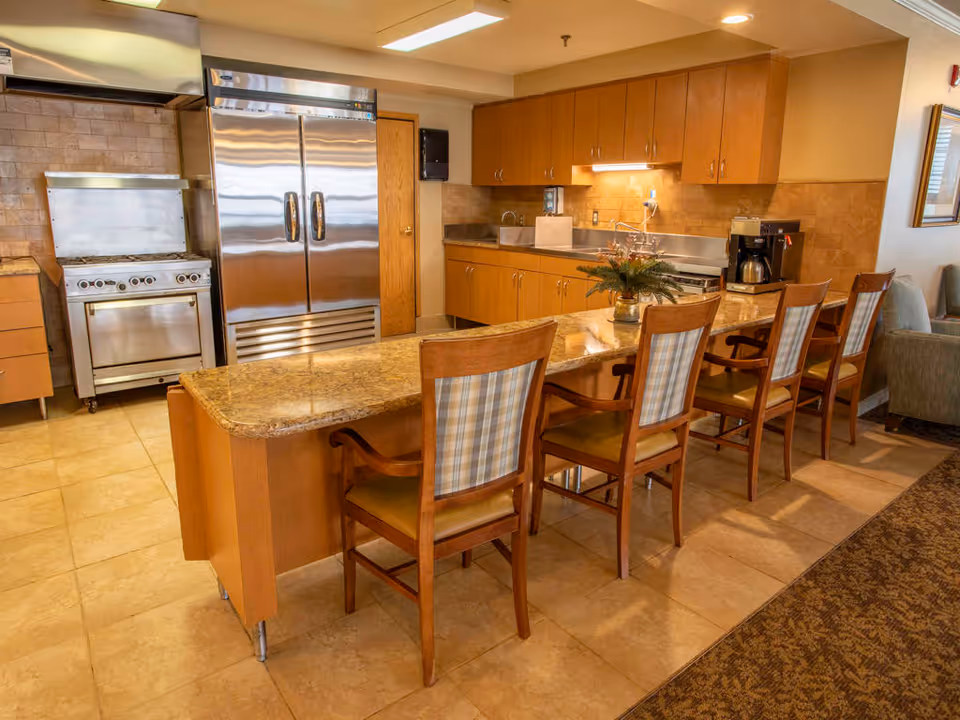 A kitchen area in a senior living facility featuring a long granite countertop with five wooden chairs lined up along one side. The kitchen has wooden cabinets, a stainless steel refrigerator, a stove, a coffee maker, and a small plant on the counter. The floor is tiled, and the lighting is warm and inviting.