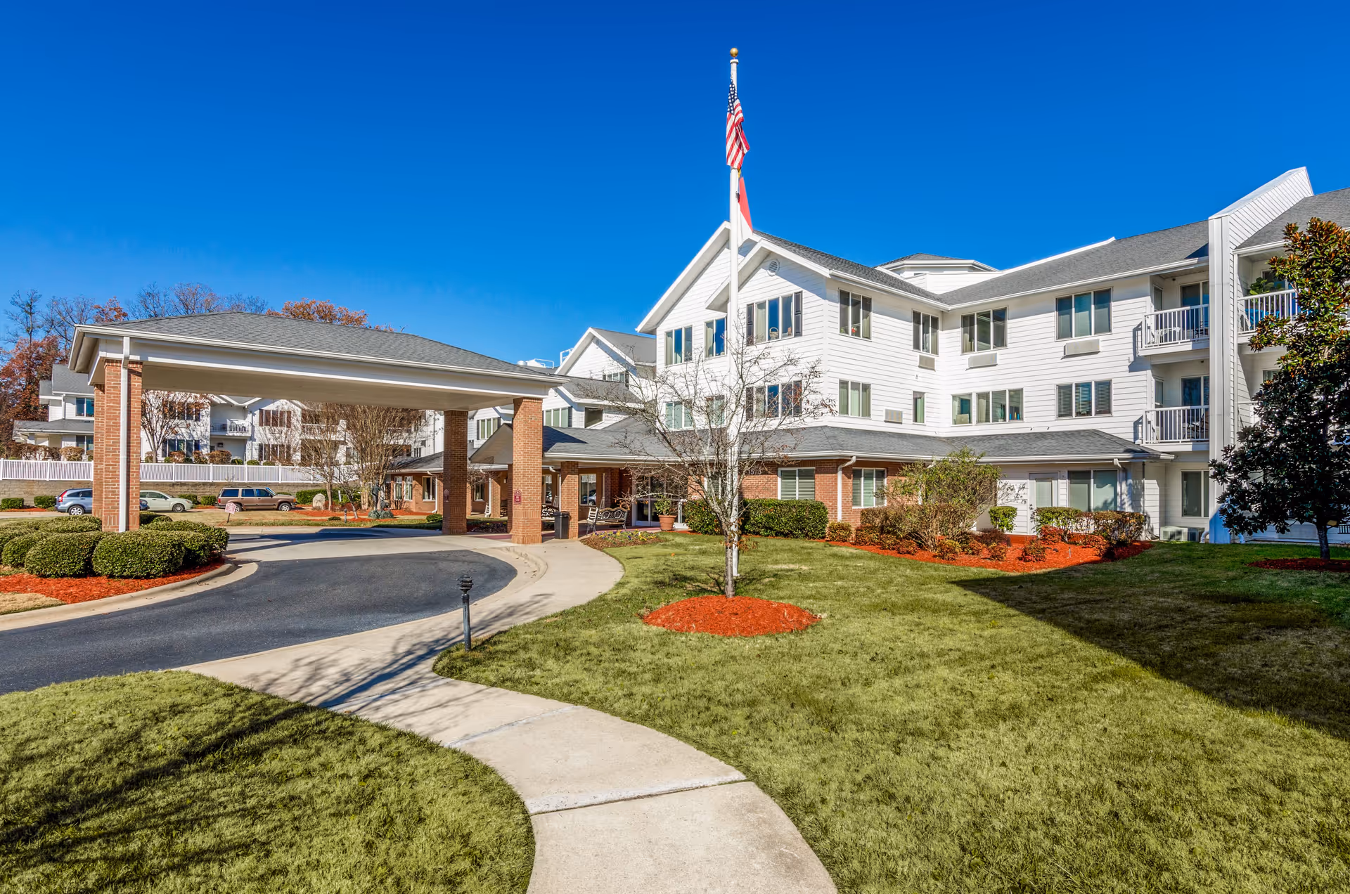 Senior living facility exterior with curved driveway, flagpole, and well-kept landscaping.