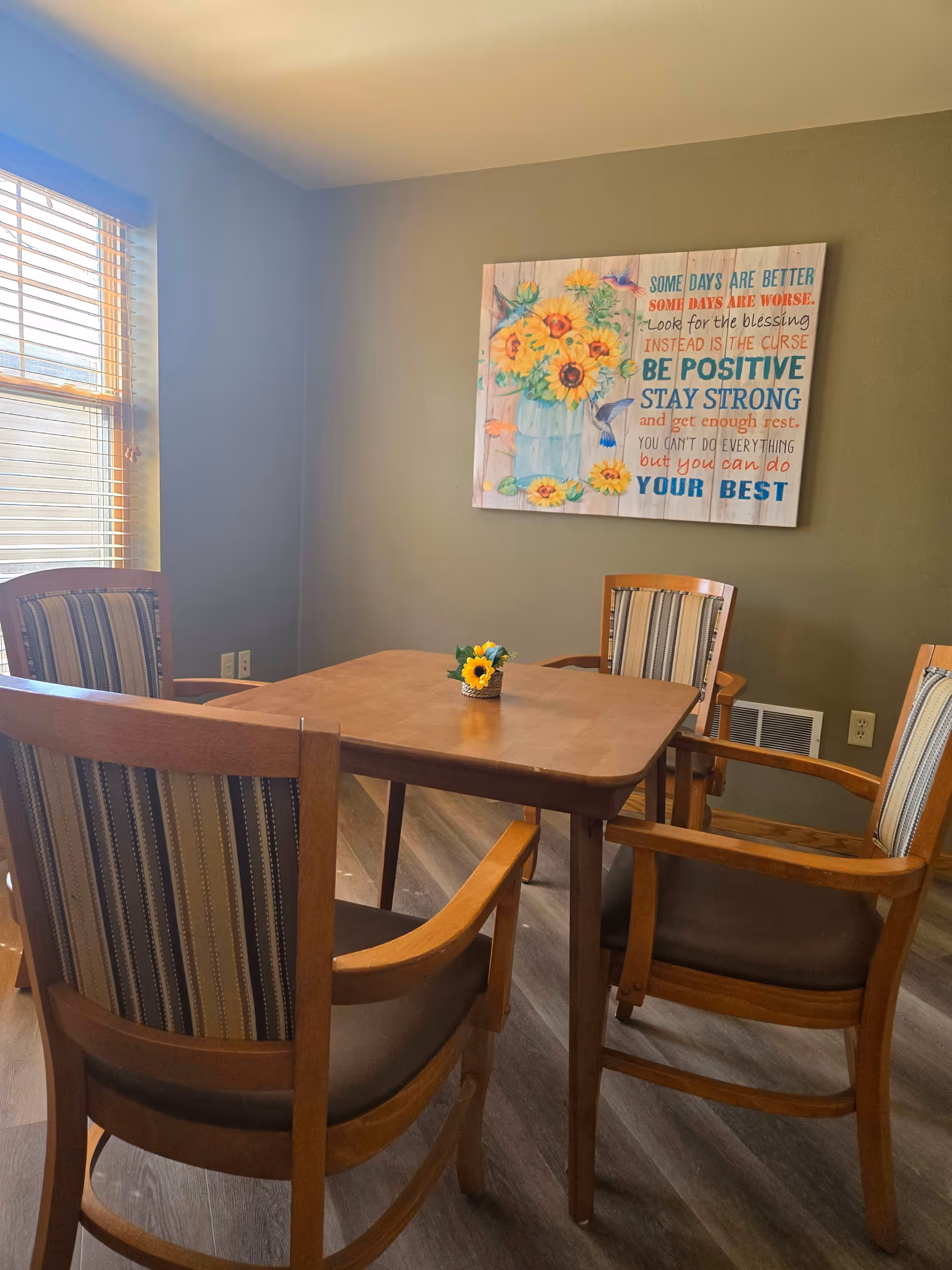 A small dining area with a wooden table and four wooden chairs with striped cushions. A small sunflower decoration is placed in the center of the table. On the wall behind the table is a colorful framed artwork featuring sunflowers and an inspirational quote. A window with blinds allows natural light into the room.