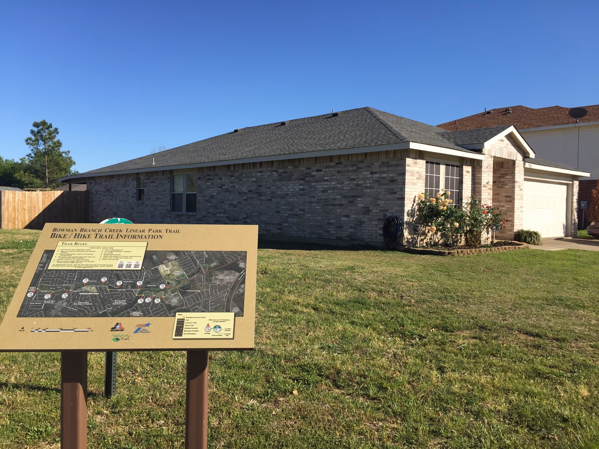 A single-story brick house with a gray shingled roof and a small garden with bushes in front. In the foreground, there is a signboard displaying a map and information about the Bowman Branch Creek Linear Park Trail for biking and hiking. The sky is clear and blue.