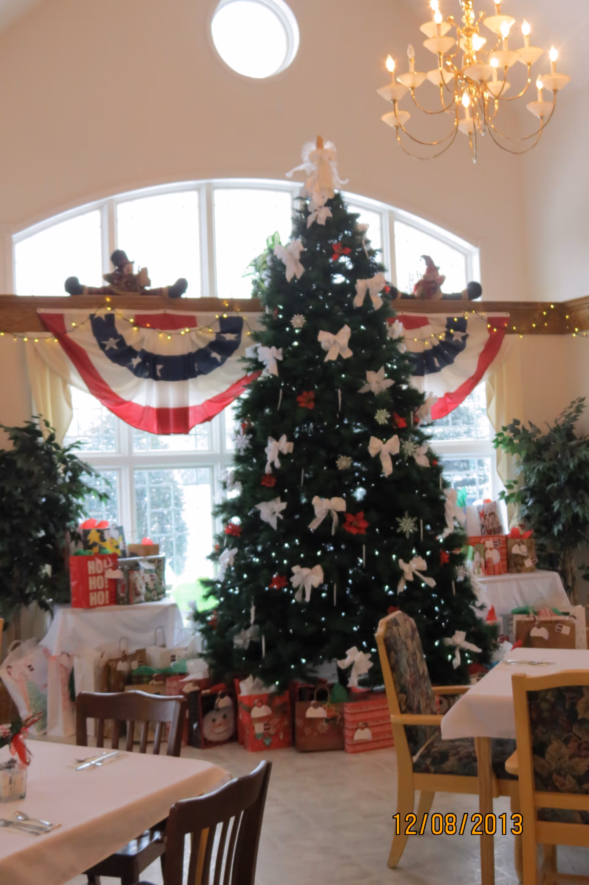 A large decorated Christmas tree with white bows and ornaments stands in a room with large windows and patriotic red, white, and blue bunting. Wrapped gifts are placed around the base of the tree. The room has tables and chairs with floral upholstery, and a chandelier hangs from the ceiling.