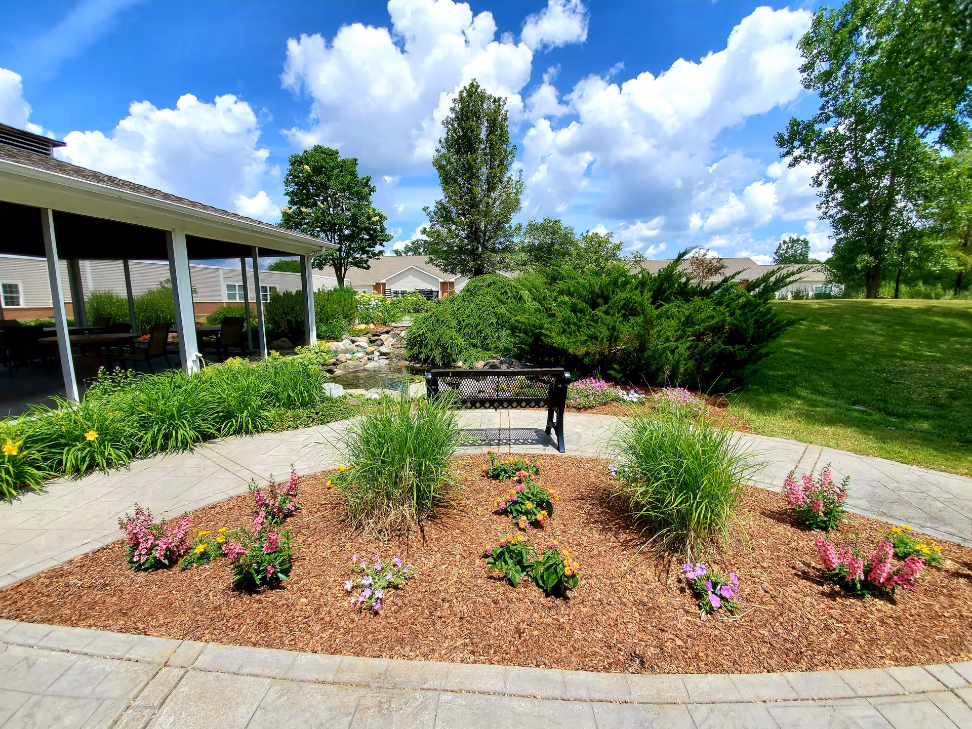 A landscaped outdoor garden area with a curved concrete pathway surrounding a mulched flower bed with various plants and flowers. There is a black metal bench facing a small pond with rocks and greenery. Trees and shrubs are visible in the background under a partly cloudy blue sky.