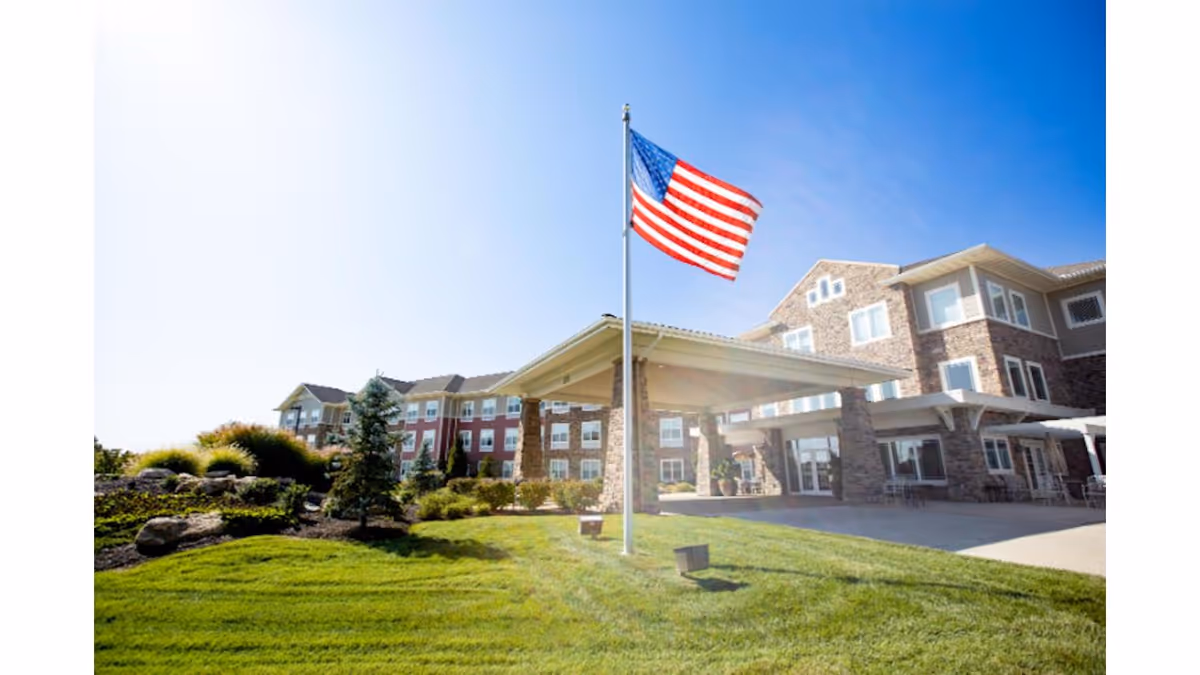 Front entrance of a multi-story brick senior living building with an American flag, covered porte-cochère, and landscaped lawn under a blue sky.