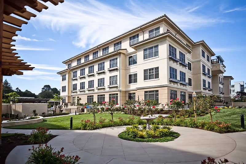 Exterior view of a multi-story senior living facility building with large windows, surrounded by a well-maintained garden with green grass, flowering plants, and a circular paved walkway with a small fountain in the center under a blue sky with some clouds.