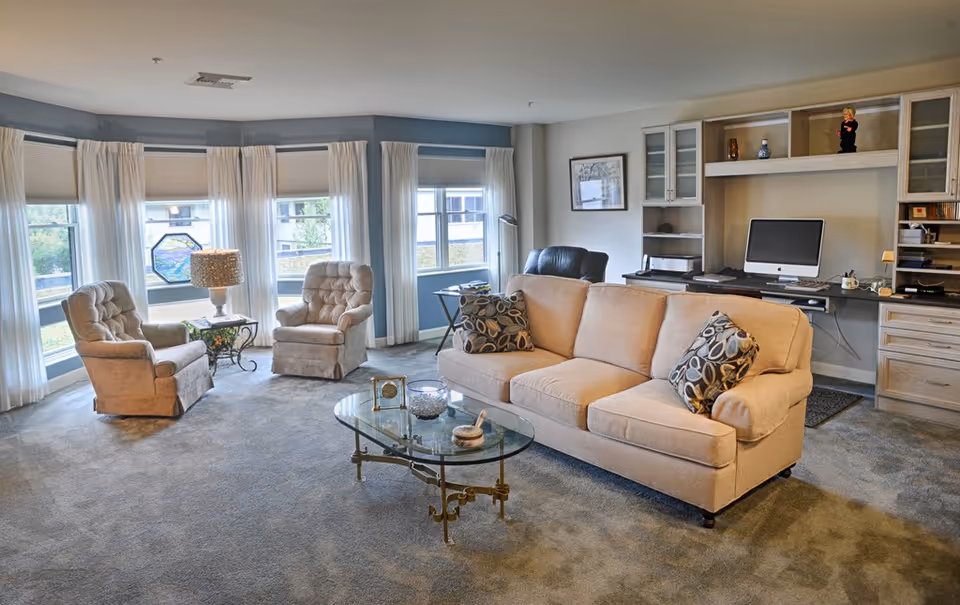 A cozy living room with a beige sofa adorned with patterned pillows, two beige armchairs near large windows with white curtains, a glass coffee table with decorative items, and a built-in desk area with a computer and shelves.