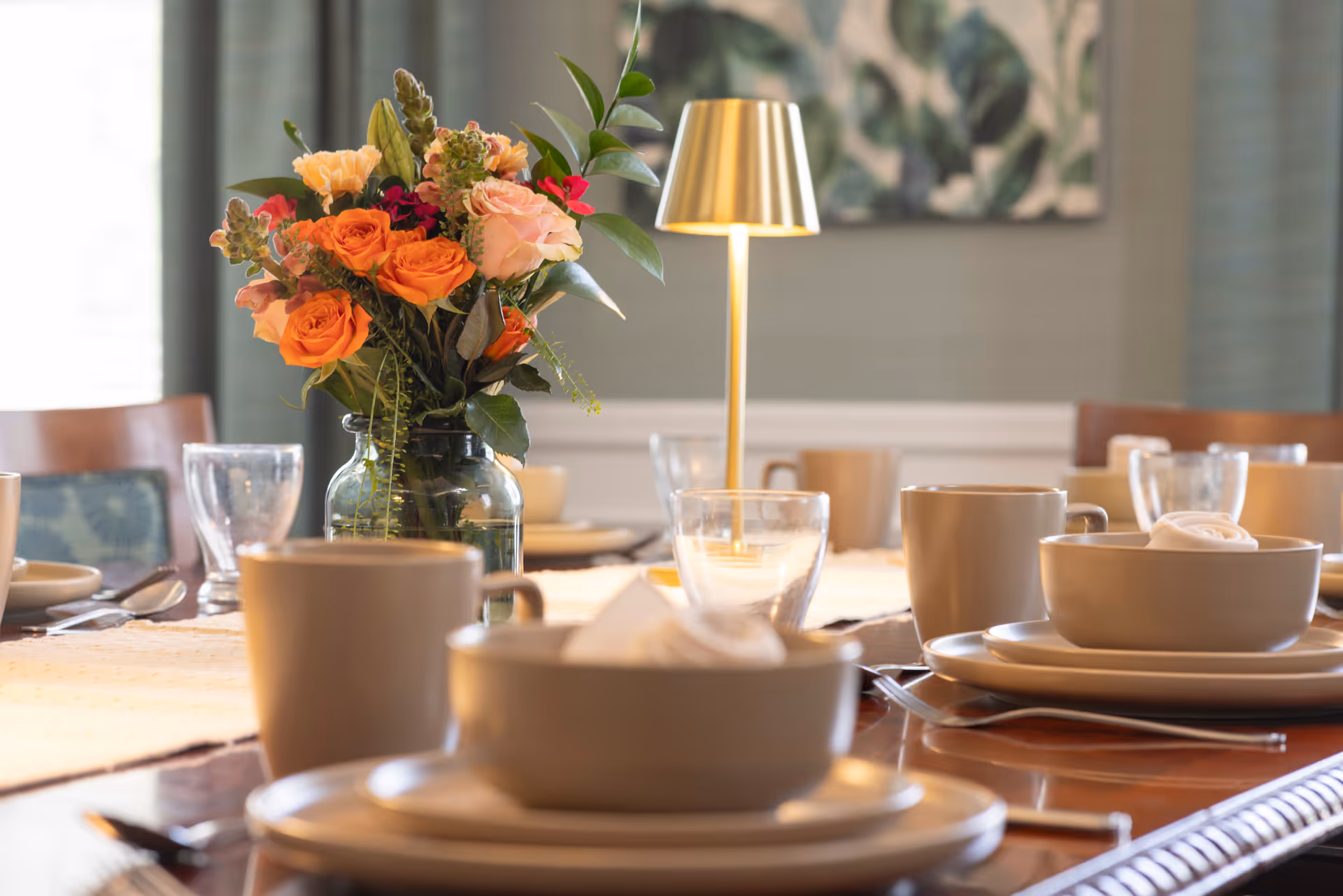 A dining table set with plates, bowls, cups, glassware, a vase of orange and pink flowers, and a small table lamp.