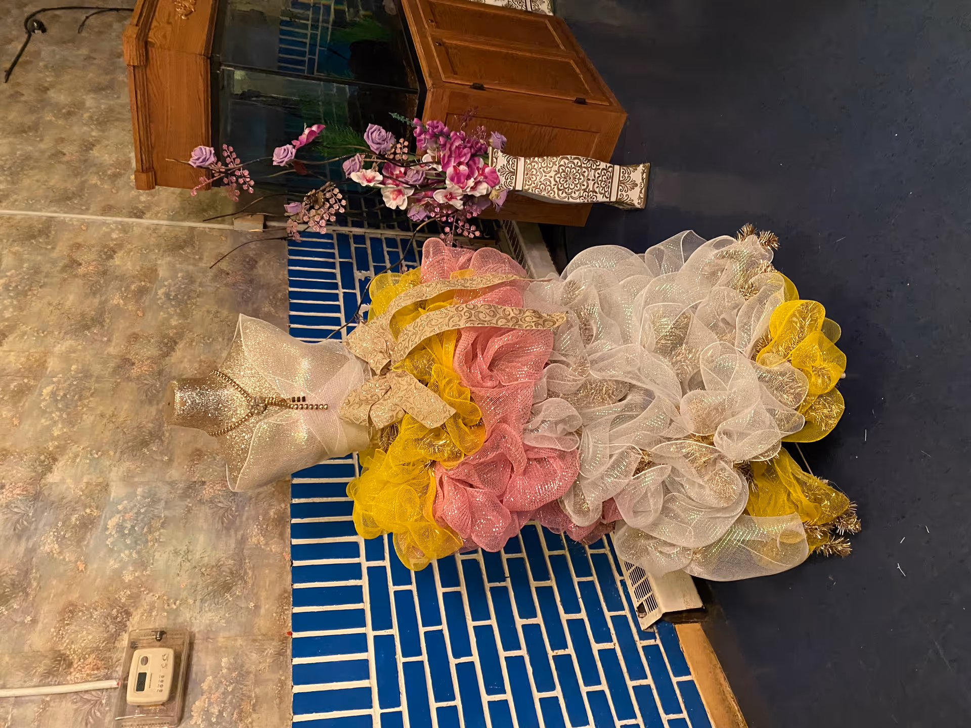 A decorative mannequin dressed in a colorful, layered dress made of yellow, pink, and white mesh fabric with a large bow at the waist, standing indoors against a blue and white tiled wall. To the right, there is a tall vase with purple and pink artificial flowers next to a wooden fish tank cabinet.