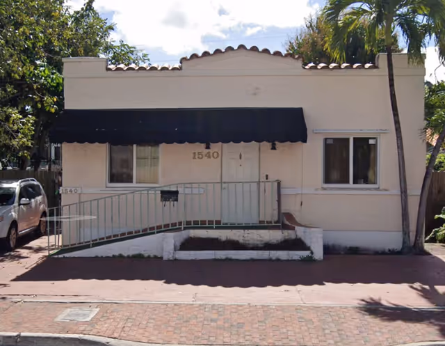 Front exterior view of a single-story building with a beige facade, a black awning above the entrance, a wheelchair ramp with green railings, and the number 1540 displayed next to the door. There is a white SUV parked on the left side and palm trees in the background.