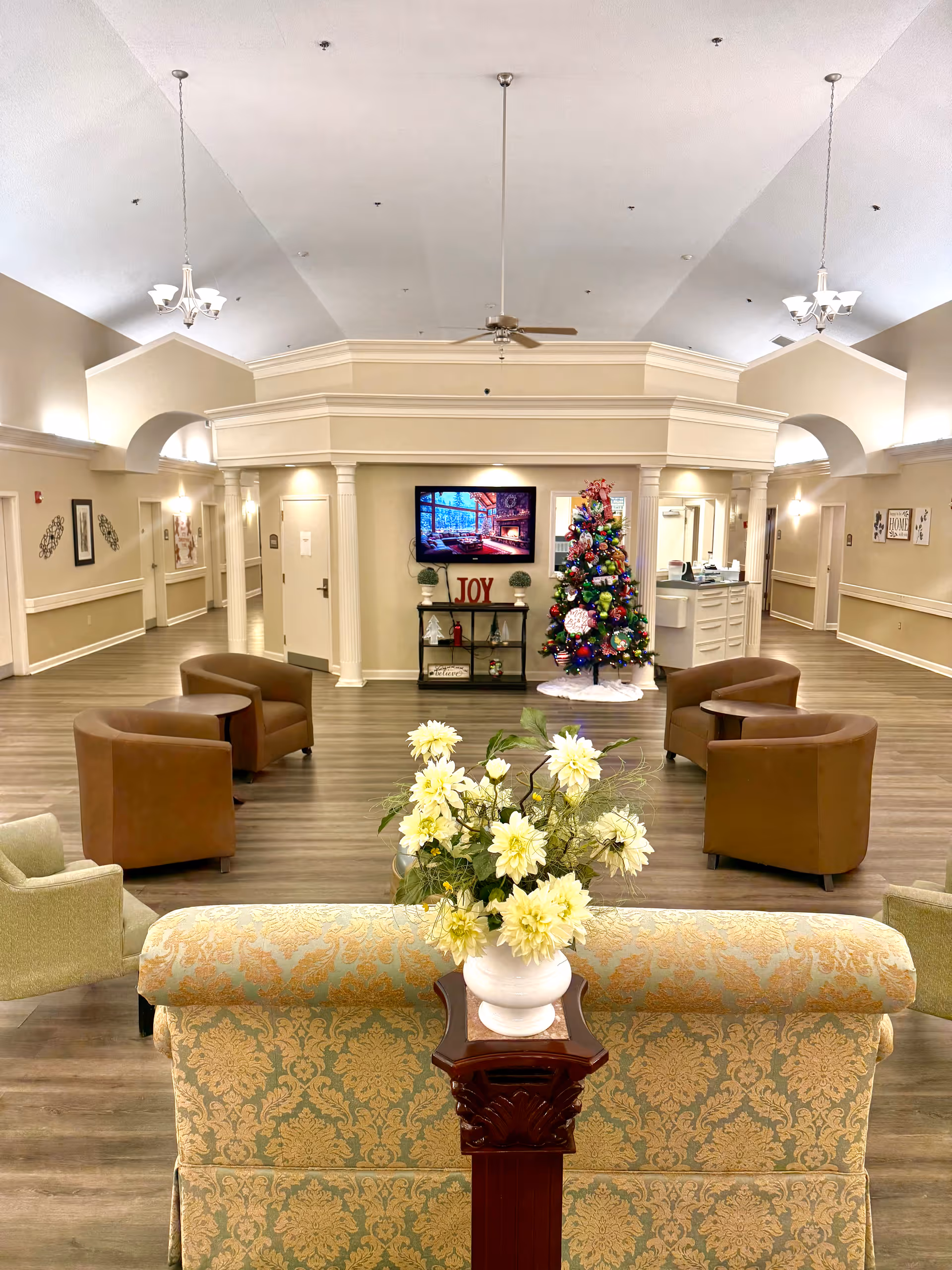 A spacious senior living facility common area with a high ceiling, beige walls, and wood flooring. The room features a decorated Christmas tree, a TV mounted on the wall above a shelf with holiday decor including a sign that says 'JOY'. There are several comfortable chairs arranged in small groups and a floral arrangement on a wooden pedestal in the foreground. The area is well-lit with hanging light fixtures and wall sconces.