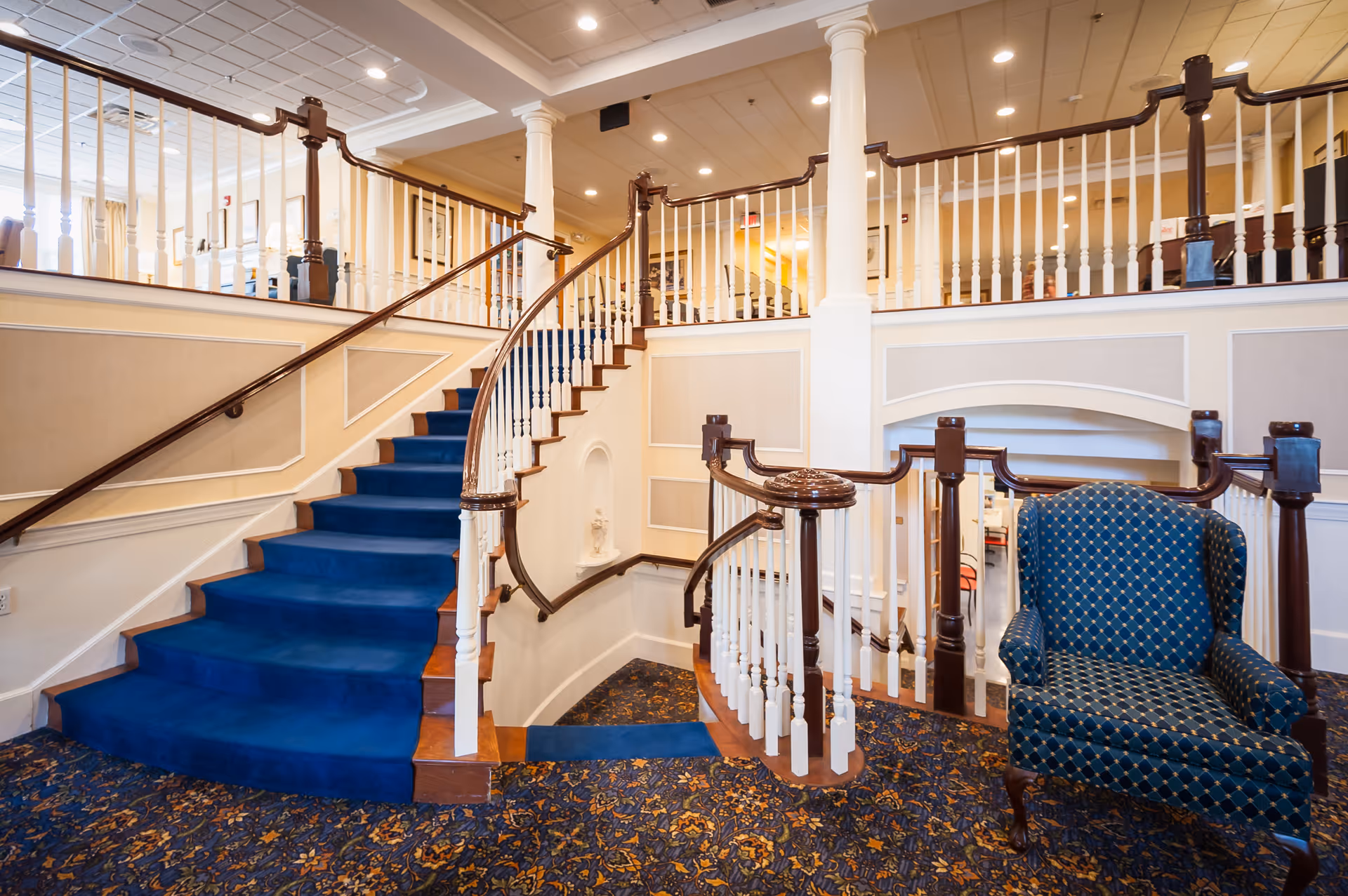 Interior view of a senior living facility featuring a curved staircase with blue carpet, white railings with dark wooden handrails, beige walls with white trim, and a blue patterned armchair on a floral carpeted floor.