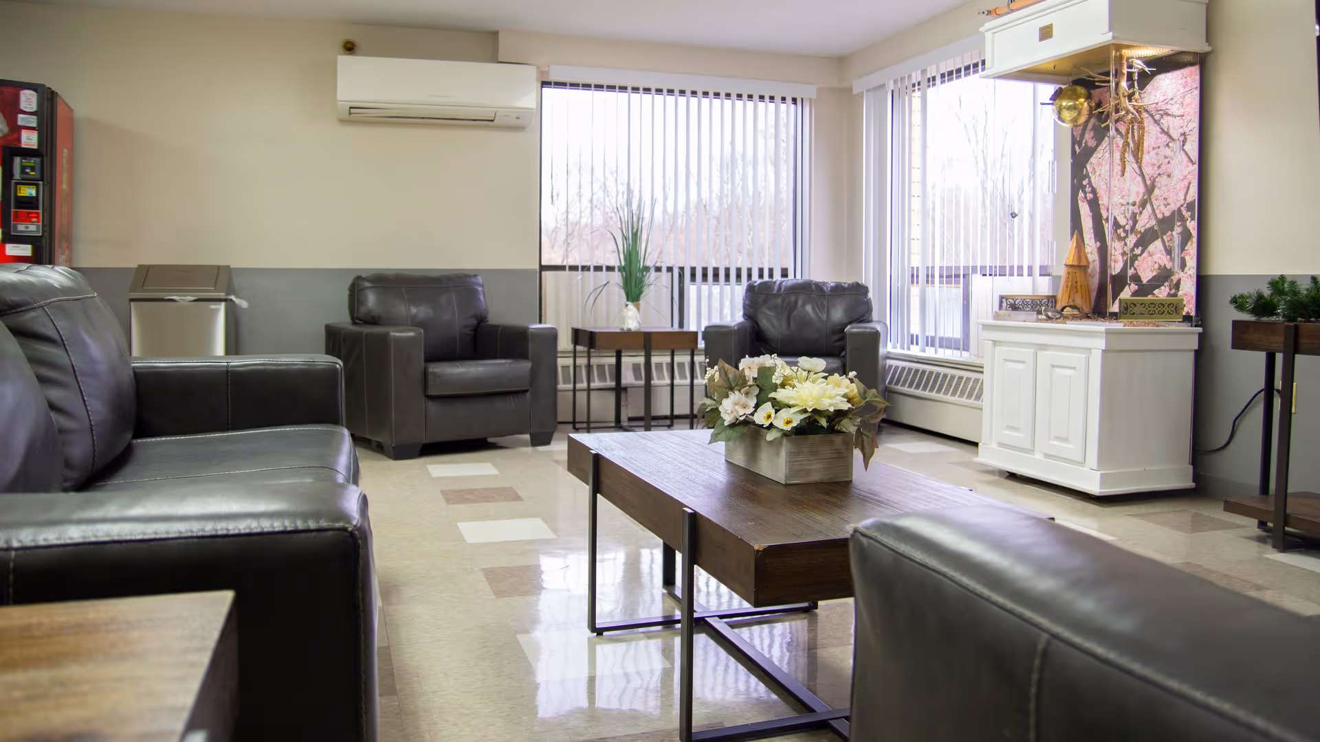 A bright and clean common area with leather armchairs and sofas arranged around a wooden coffee table with a floral centerpiece. Large windows with vertical blinds allow natural light to fill the room. There is an air conditioning unit mounted on the wall and a vending machine in the corner.