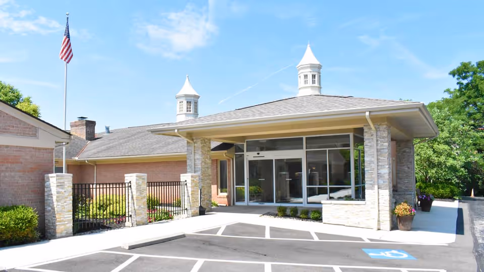 Front exterior view of Willow Brook Christian Home showing a single-story building with a covered entrance supported by stone pillars, two cupolas on the roof, an American flag on a flagpole, and a parking area with a handicapped parking space.