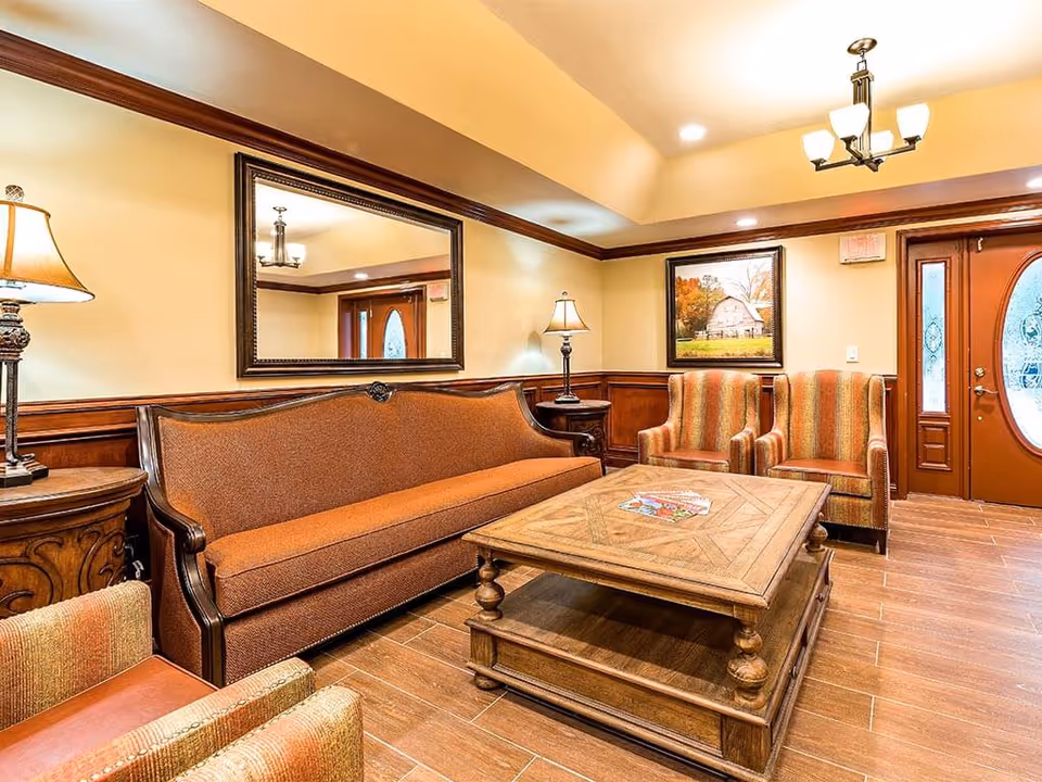 A cozy senior living facility common area with a long brown upholstered sofa, two striped armchairs, a wooden coffee table with magazines, two side tables with lamps, a large mirror on the wall, a framed picture of a barn, and a wooden door with an oval glass window.