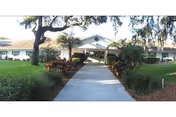 A paved walkway leading to the covered entrance of a one-story building surrounded by grass, trees, and landscaping.
