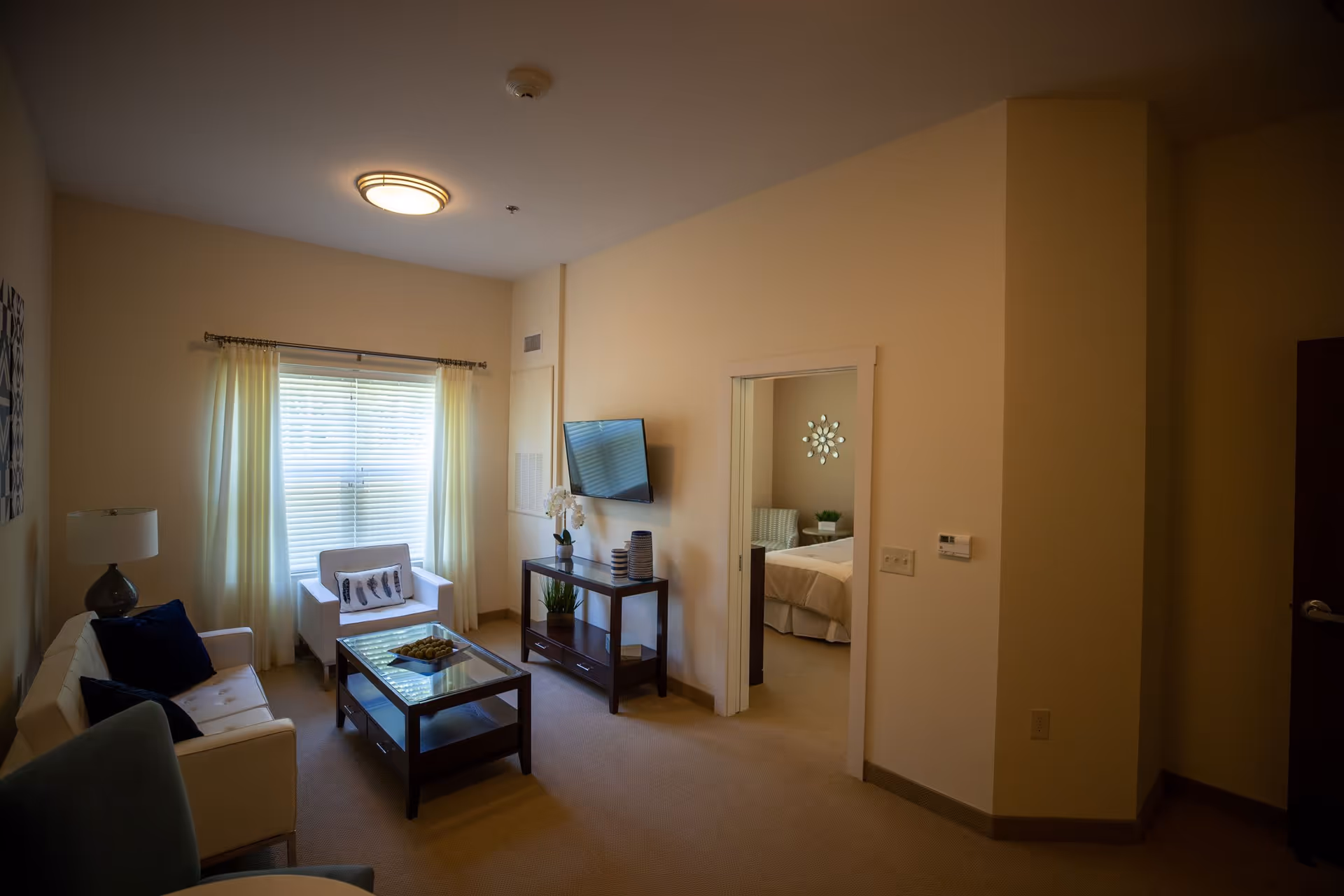 A cozy living room with a white sofa, a white armchair, a glass coffee table, and a TV mounted on the wall. The room has beige walls and carpet, a window with white curtains, and a doorway leading to a bedroom with a bed and decorative wall art.