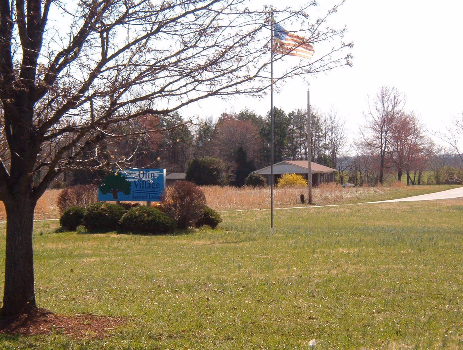 Grassy front lawn with a tree, an American flag, and a sign for Olin Village near a driveway and a small building in the background.