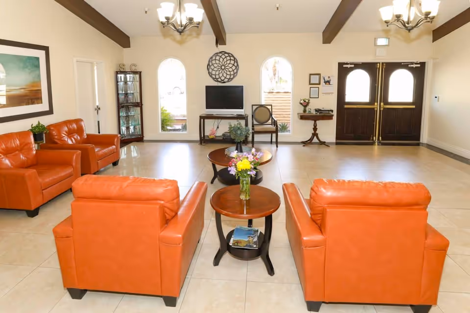 Bright assisted-living common room with four orange leather armchairs arranged around round coffee tables, a TV, decorative windows, and double entrance doors.
