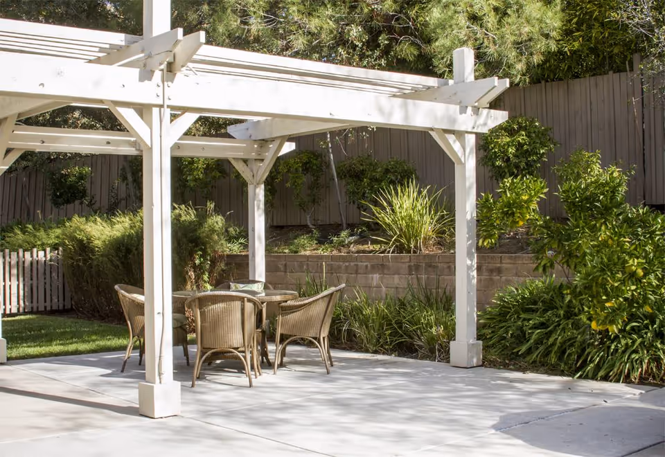 Outdoor patio area with a white wooden pergola providing partial shade over a round glass table surrounded by four wicker chairs. The patio is surrounded by green shrubs, plants, and a wooden fence in the background.