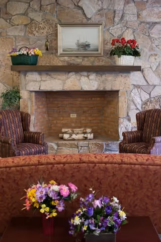 Cozy seating area with a stone fireplace flanked by two armchairs, a mantel with decor and several floral arrangements.