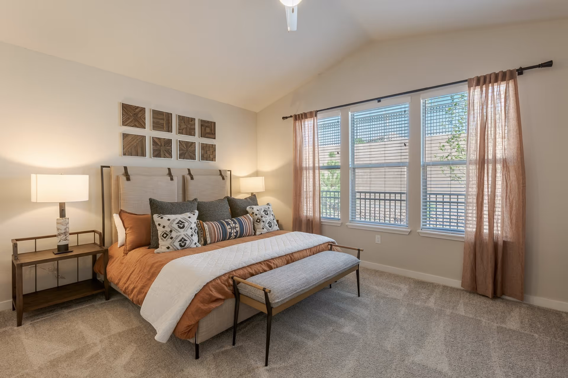 A bright bedroom with a large bed featuring multiple decorative pillows and a rust-colored bedspread with a white quilted throw. There are two bedside tables each with a lamp, a bench at the foot of the bed, and three large windows with sheer rust-colored curtains letting in natural light. The walls are light-colored with a set of eight wooden square art pieces above the bed.