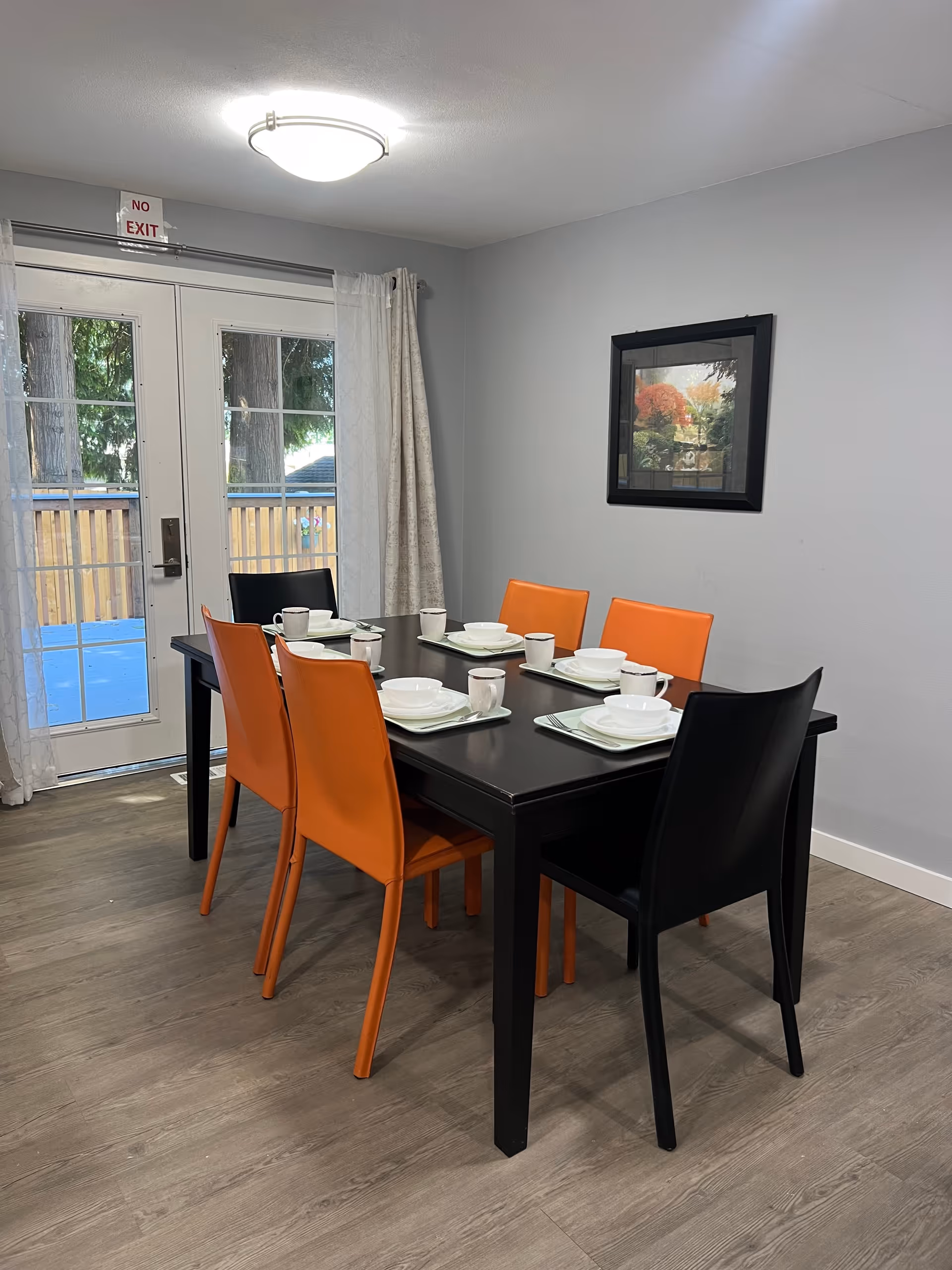A dining room with a black rectangular table set for six people. The table has white plates, bowls, cups, and napkins on trays. There are six chairs around the table, four orange and two black. The room has light gray walls, a framed picture on the wall, and double glass doors with sheer curtains leading outside. A ceiling light fixture is visible above the table.