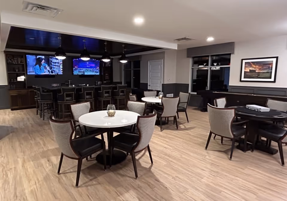 A modern dining and lounge area with round tables and chairs arranged on a light wood floor. In the background, there is a bar with high stools and two large wall-mounted televisions displaying different programs. The room has recessed lighting and a framed picture on the wall near a window.