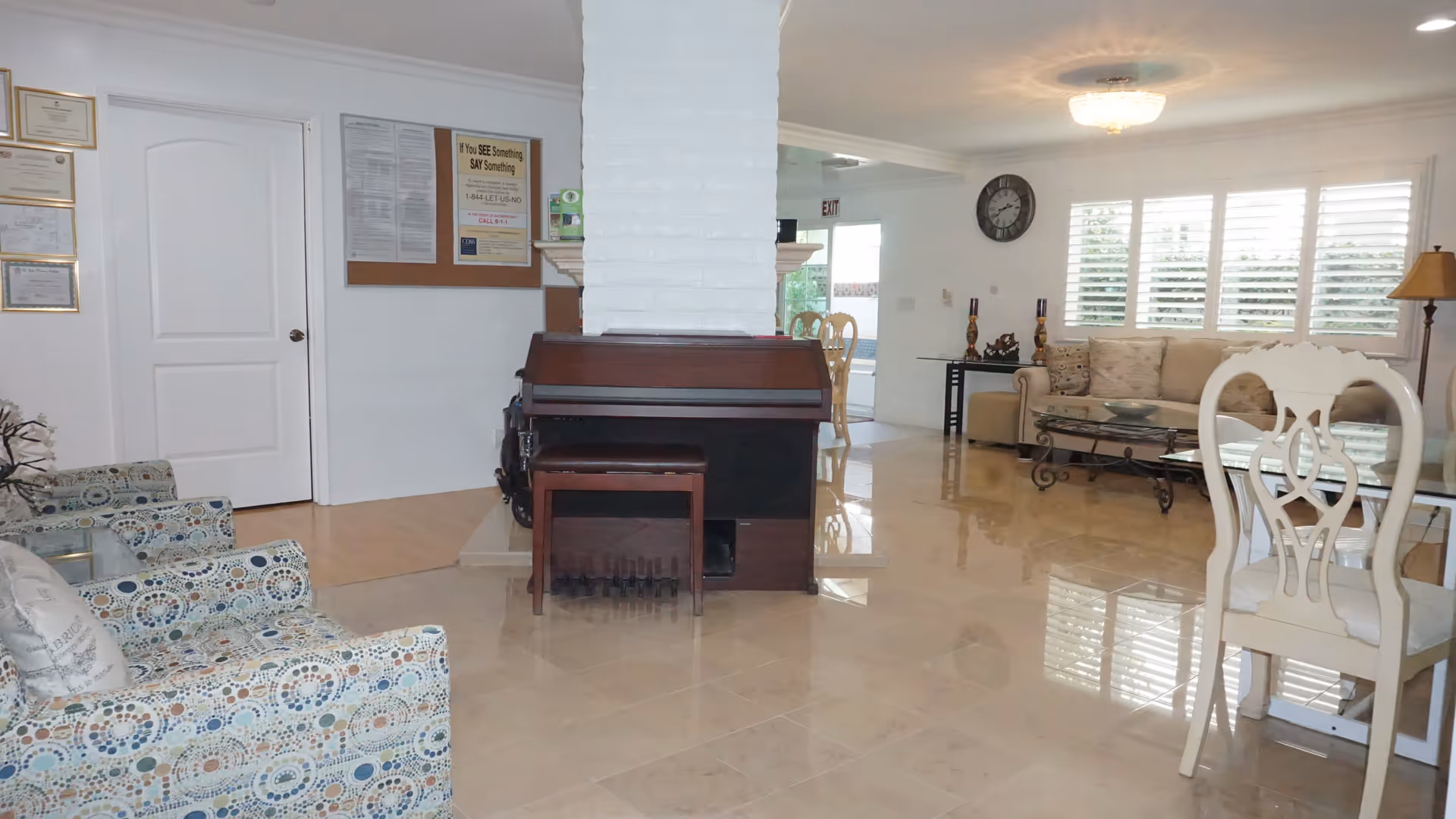 Interior view of a living room area in Olive Branch Assisted Living featuring a piano with a bench in front of a white brick column, patterned armchairs, a beige sofa with cushions, a glass coffee table, a glass dining table with white chairs, a wall clock, and a floor lamp. The room has tiled floors and large windows with white shutters allowing natural light to enter.