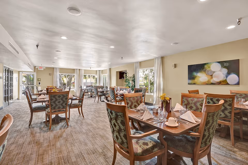 Bright communal dining room with multiple set wooden tables and upholstered chairs in a senior living facility.